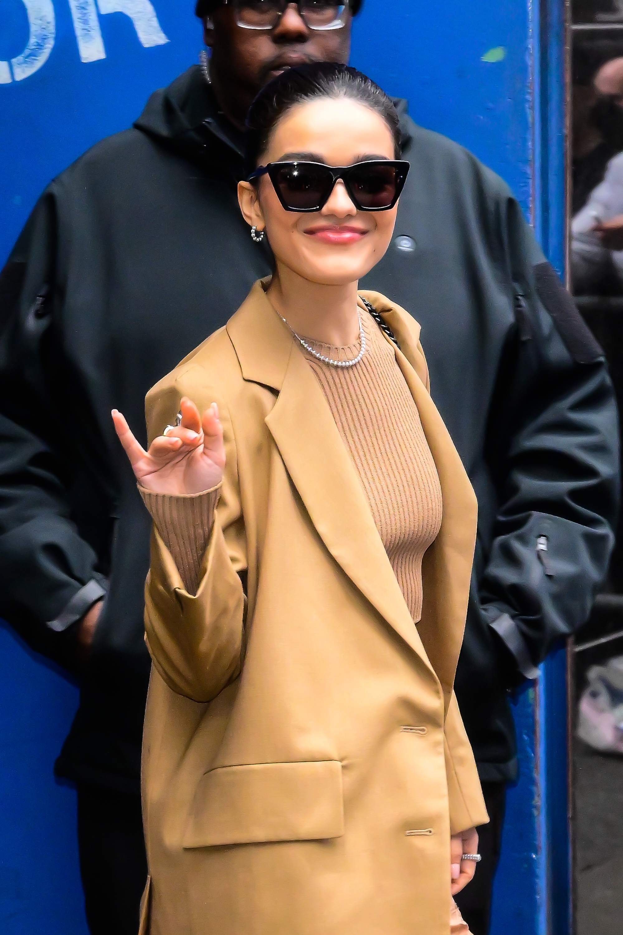 Rachel Zegler in chic sunglasses and a stylish oversized blazer smiles and waves at a public event