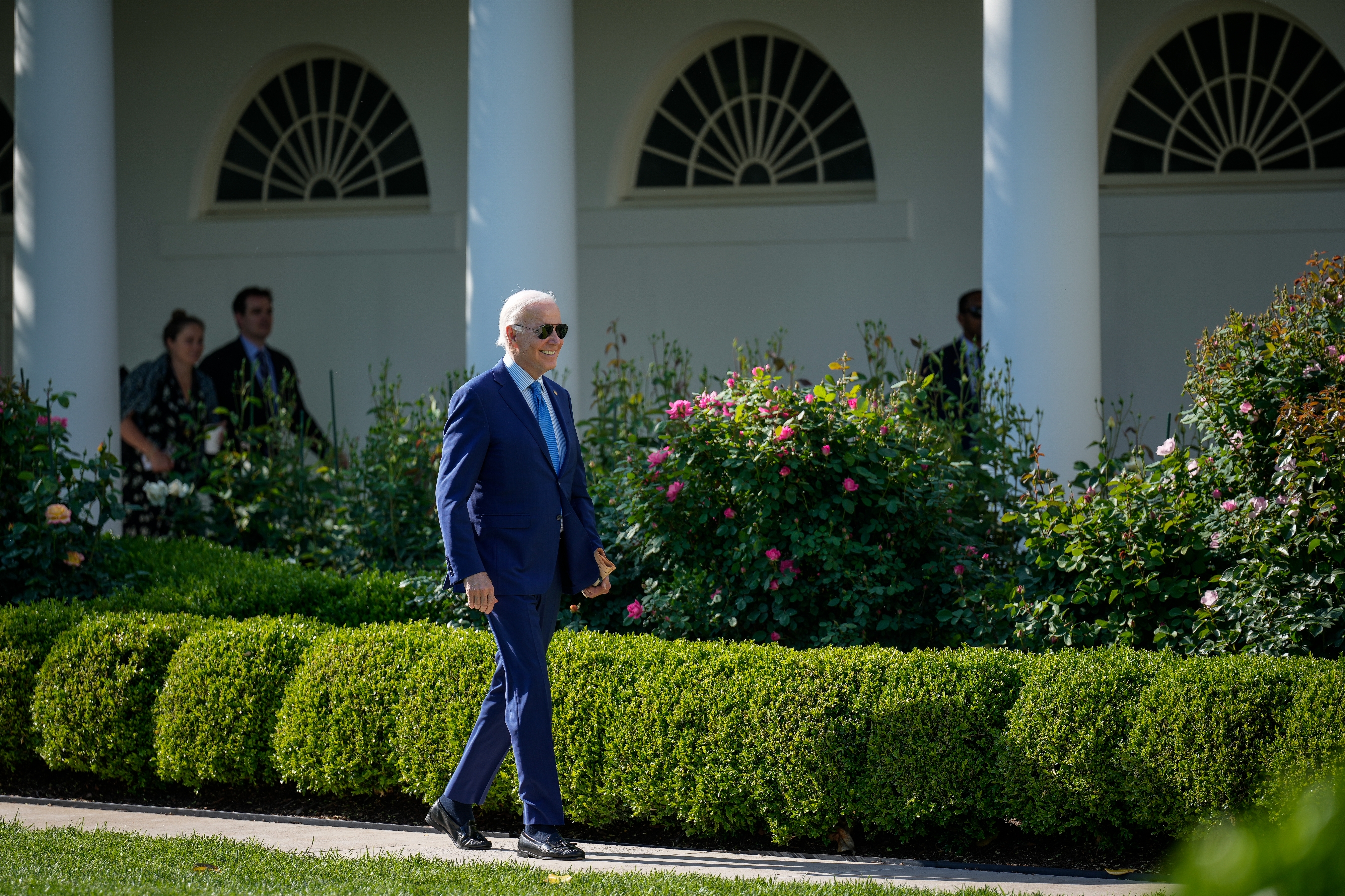 A person in a suit walks through a garden area with shrubbery and flowers, in front of a building with white columns
