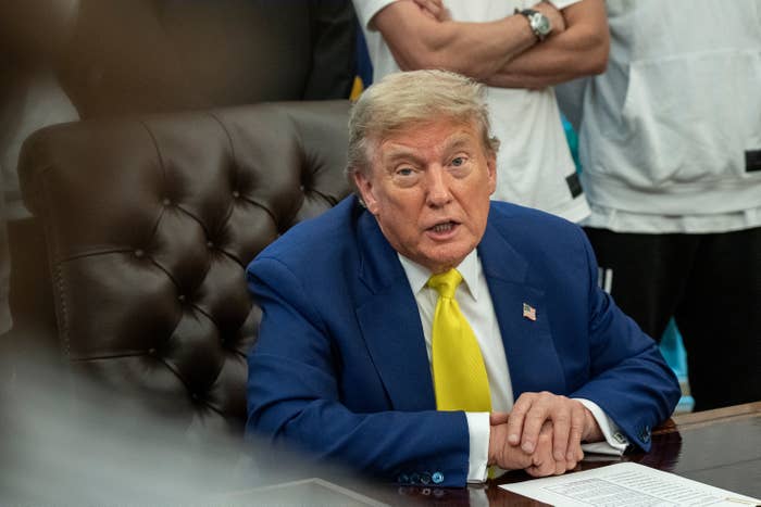 Person in a formal suit sits at a desk, speaking. Others stand in the background