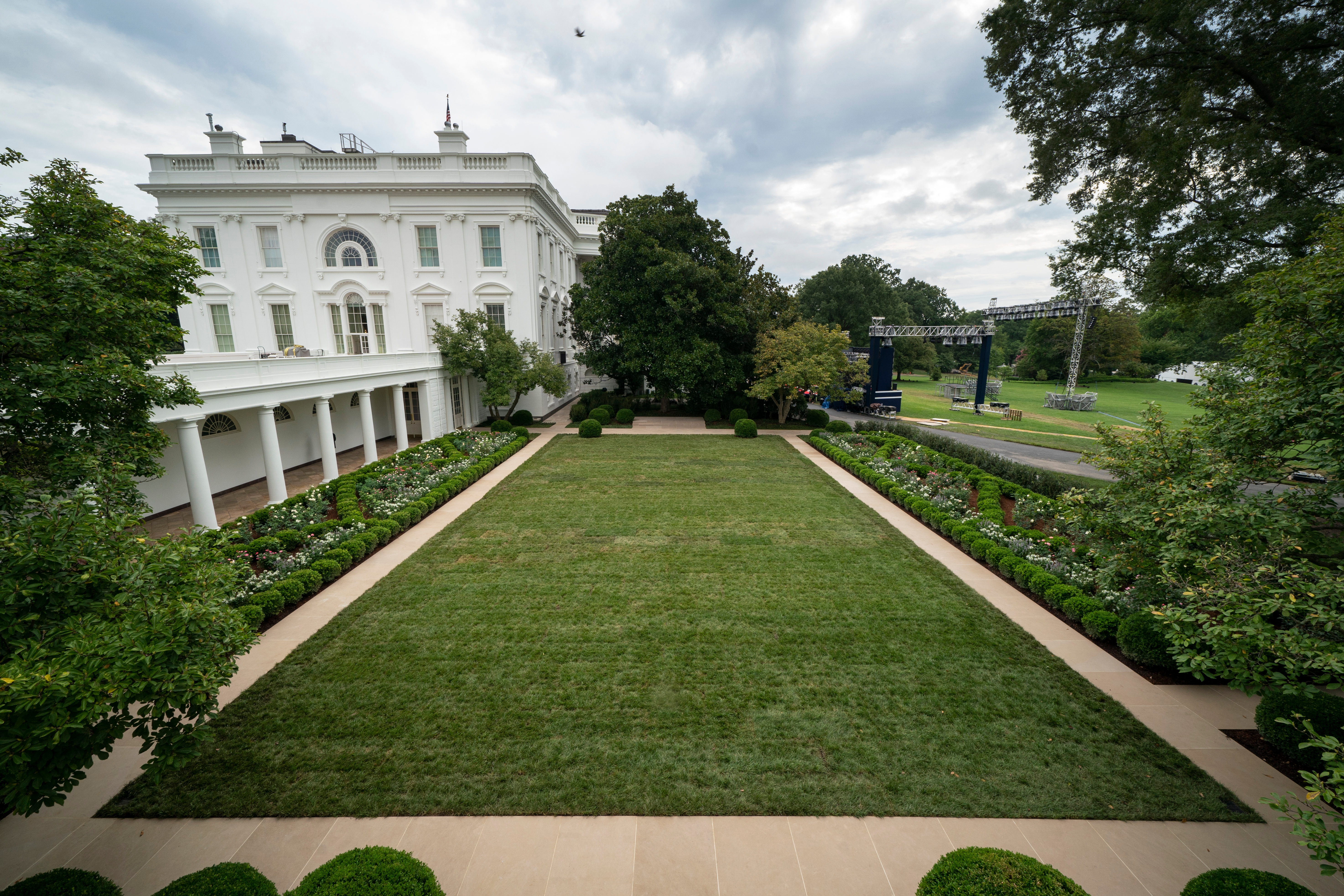 White House garden with trimmed lawn, flanked by trees and bushes, with a view of the building and a cloudy sky