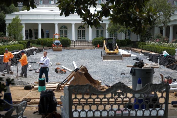 People working on a construction project in a garden area outside a large white building with columns. Machinery and materials are visible