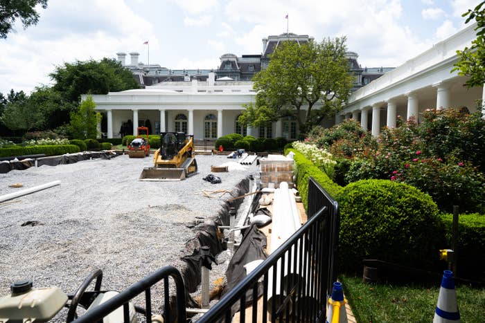 Construction scene in front of a white building with heavy machinery and gravel, surrounded by greenery and barriers
