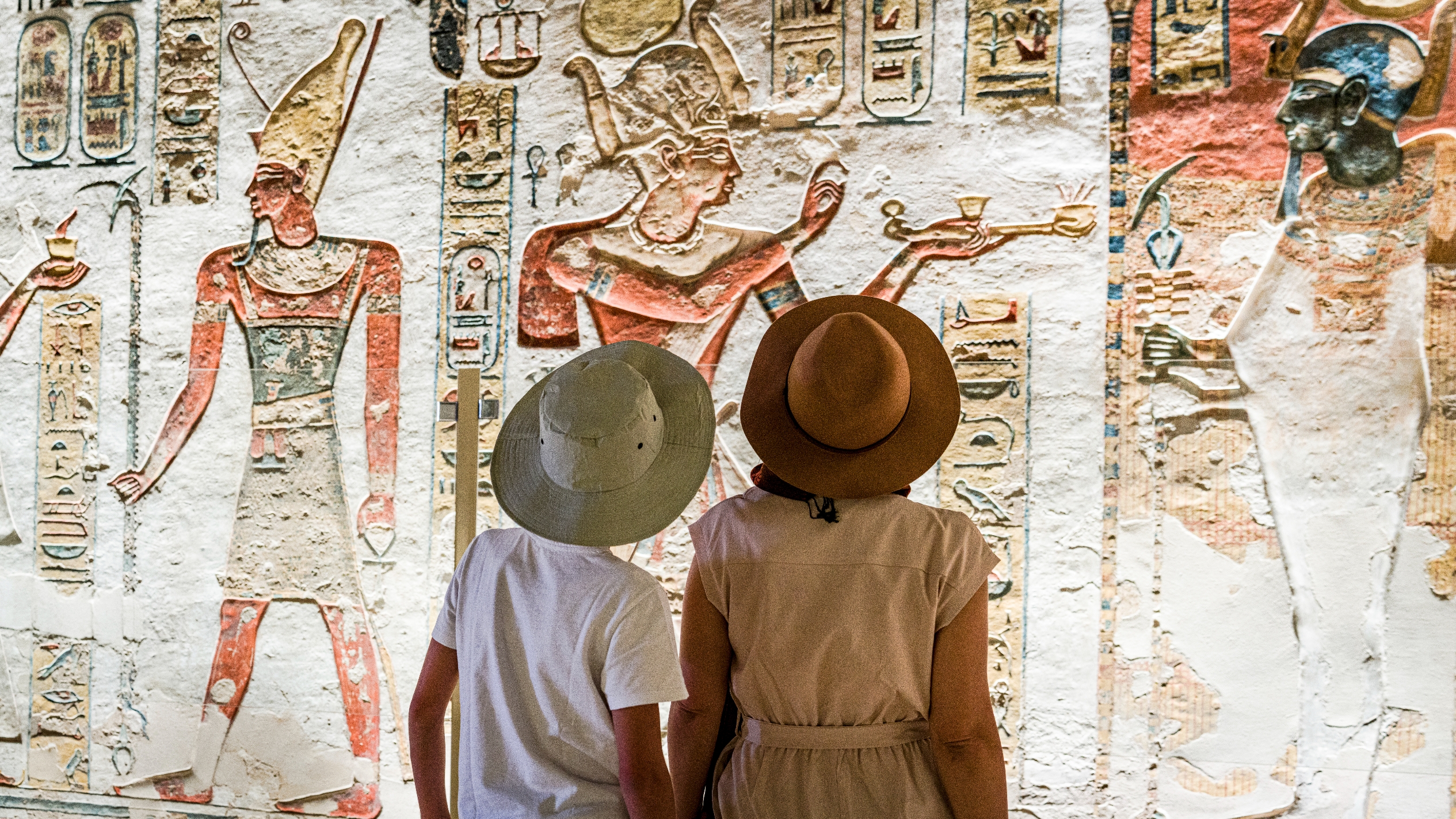 To personer i hatter beundrer eldgamle egyptiske veggkunst med detaljerte hieroglyfer og figurer i tradisjonell antrekk