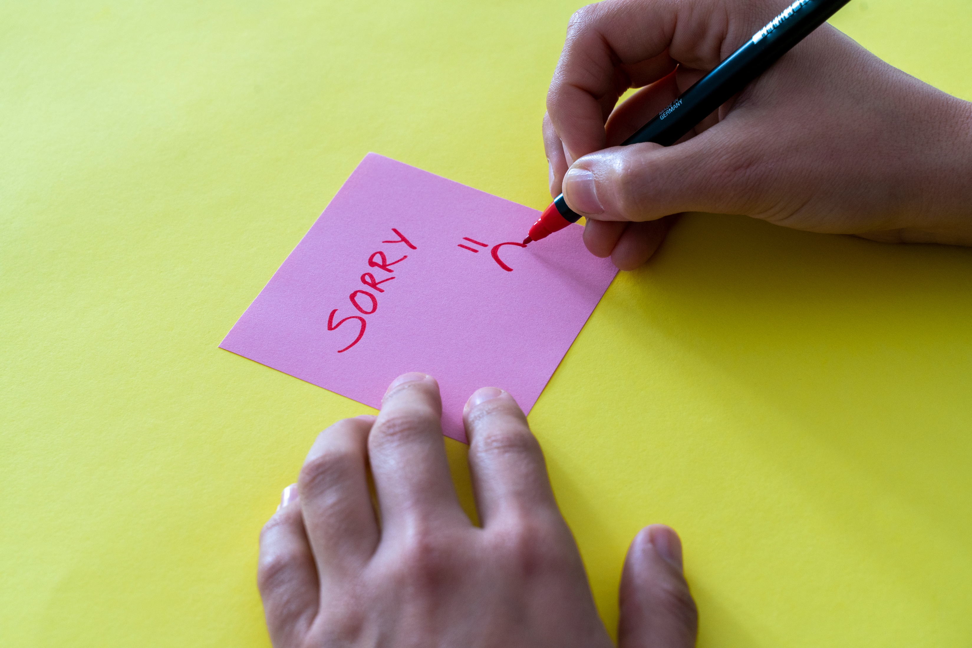 Hands Writing & quot; Beklager & quot; med et trist ansikt på en klissete lapp mot en gul bakgrunn