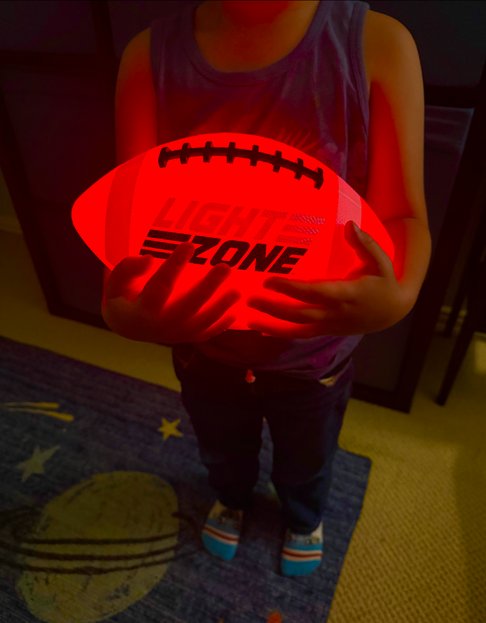 Child holding a glowing football labeled "LIGHT ZONE" indoors, wearing a sleeveless top and jeans, standing on a space-themed rug
