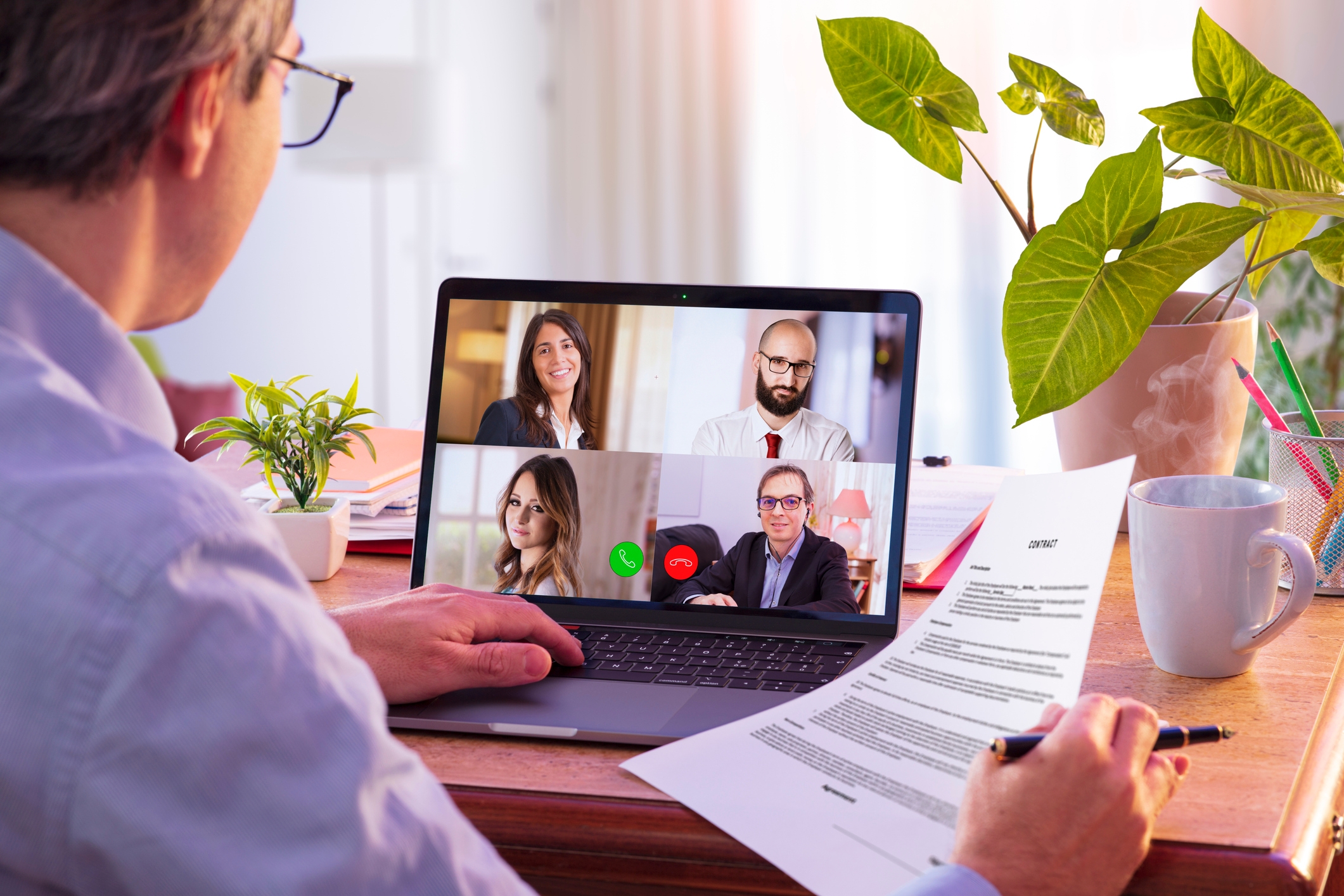 Person på videosamtaler med fire kolleger på en bærbar datamaskin, og holder et dokument ved et skrivebord med planter og et krus i nærheten