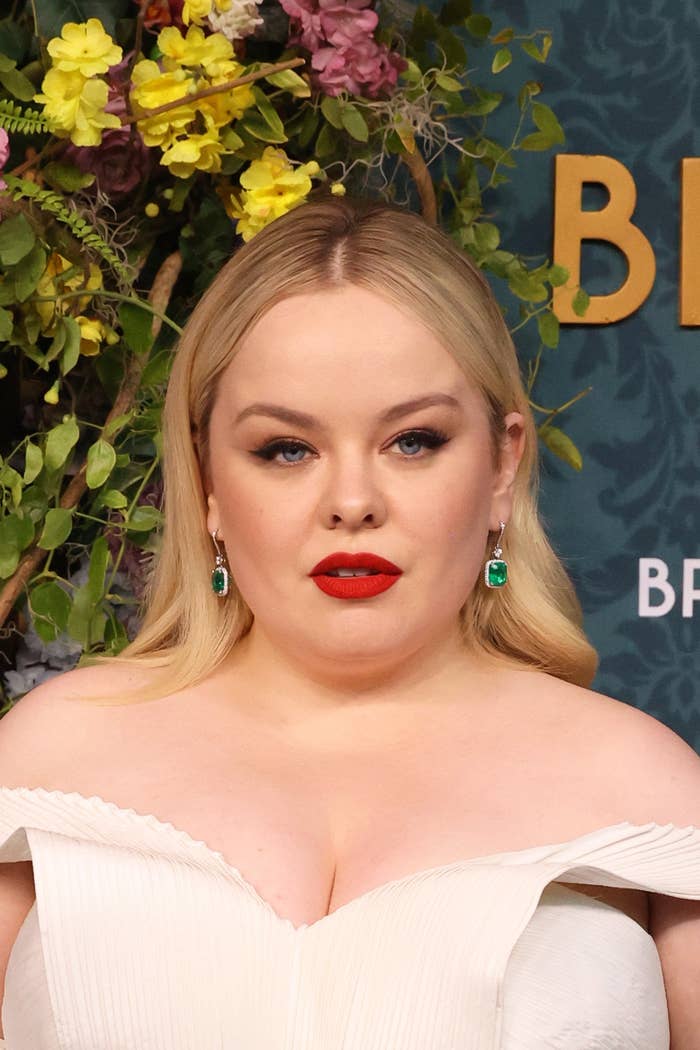 Nicola Coughlan with long hair, wearing a white off-shoulder dress, emerald earrings, and red lipstick, poses in front of a floral backdrop