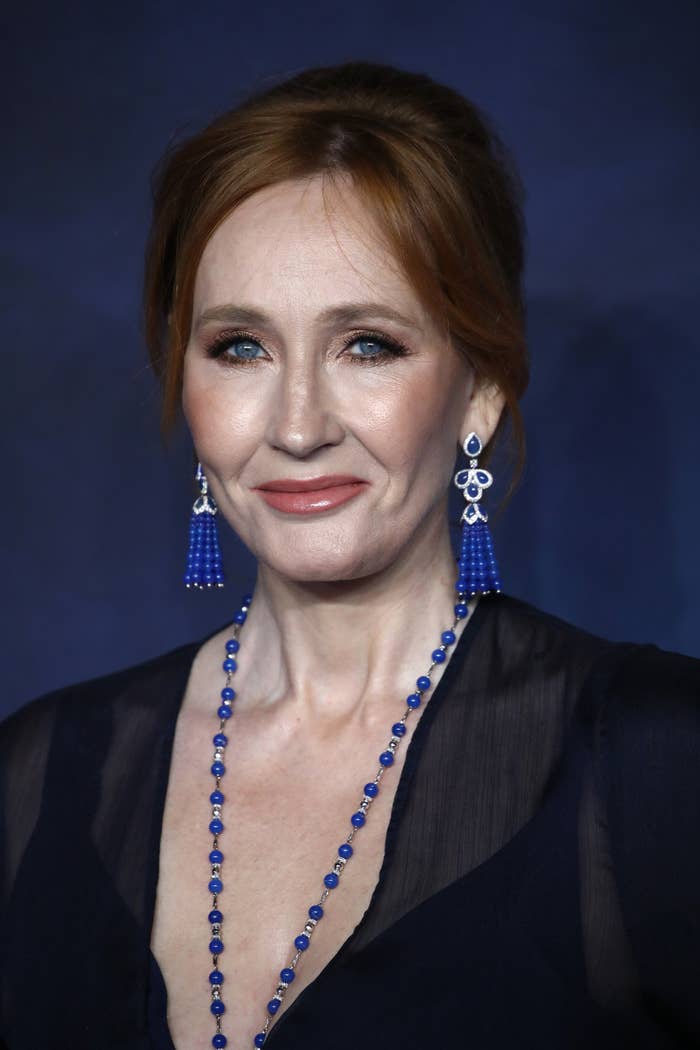 J.K. Rowling with an elegant blue necklace and drop earrings, wearing a sheer dark outfit, poses on a blue backdrop