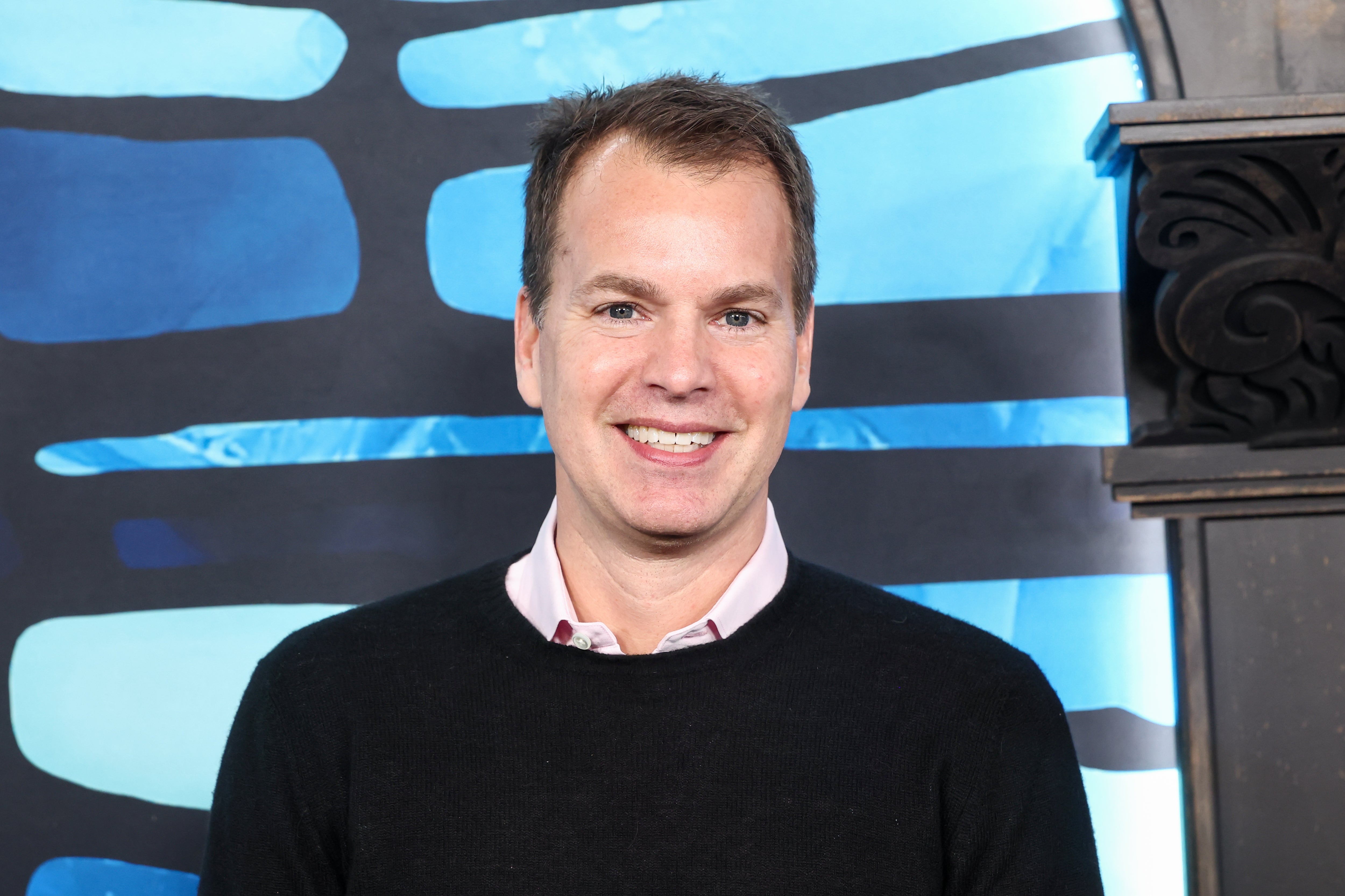 Casey Bloys smiling in a black sweater and pink shirt, standing in front of a patterned backdrop