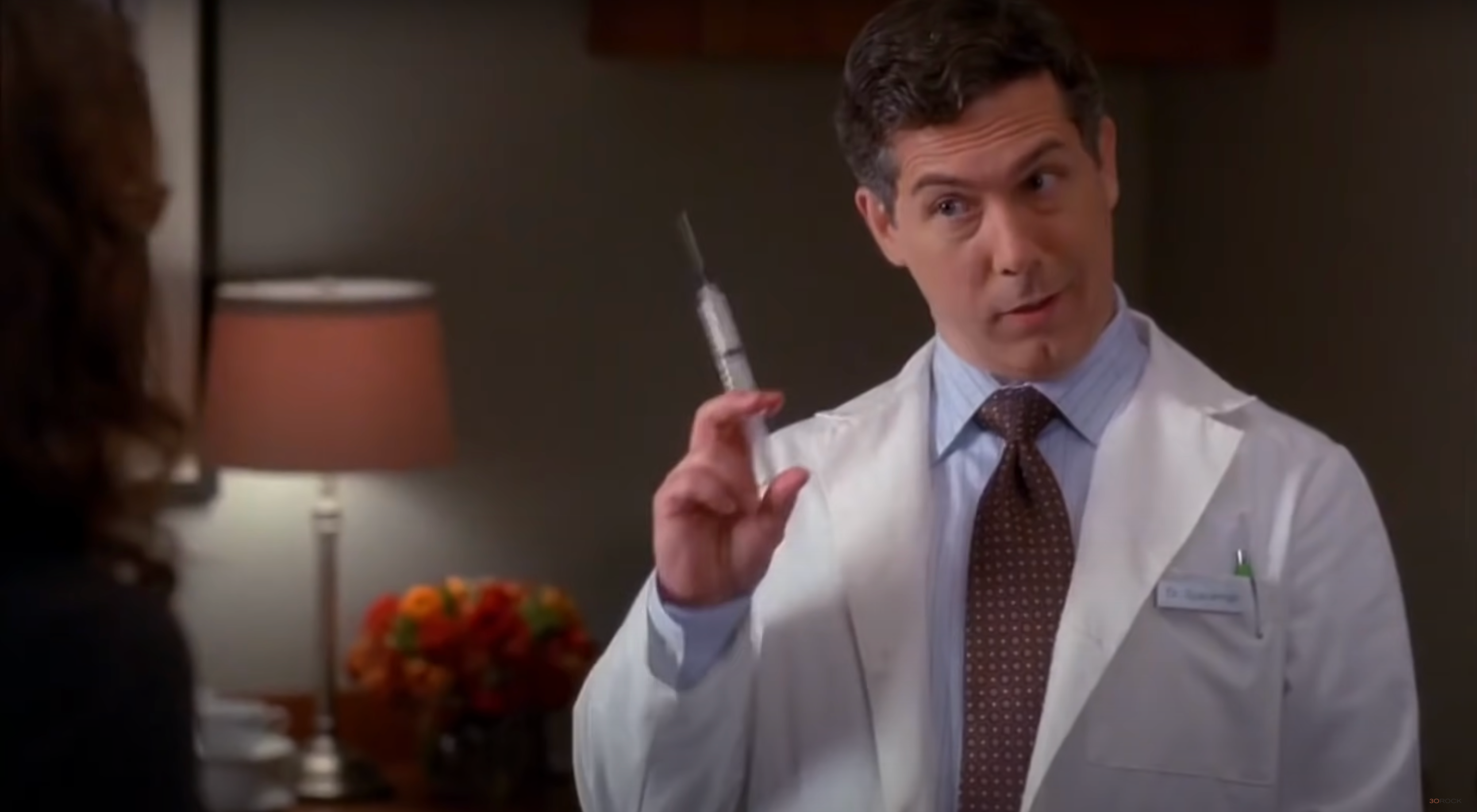 En person i en hvit lab -frakk holder en stor sprøyte, og ser ut til å snakke, i et rom med en lampe og blomster i bakgrunnen