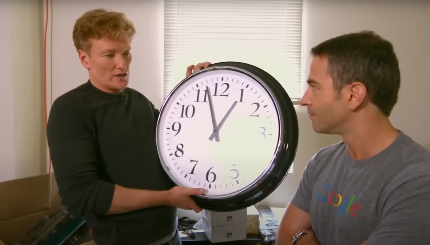 To personer innendørs, en som holder en stor klokke som viser 12:03. Den andre personen har på seg en skjorte med et gjenkjennelig teknisk selskaps logo