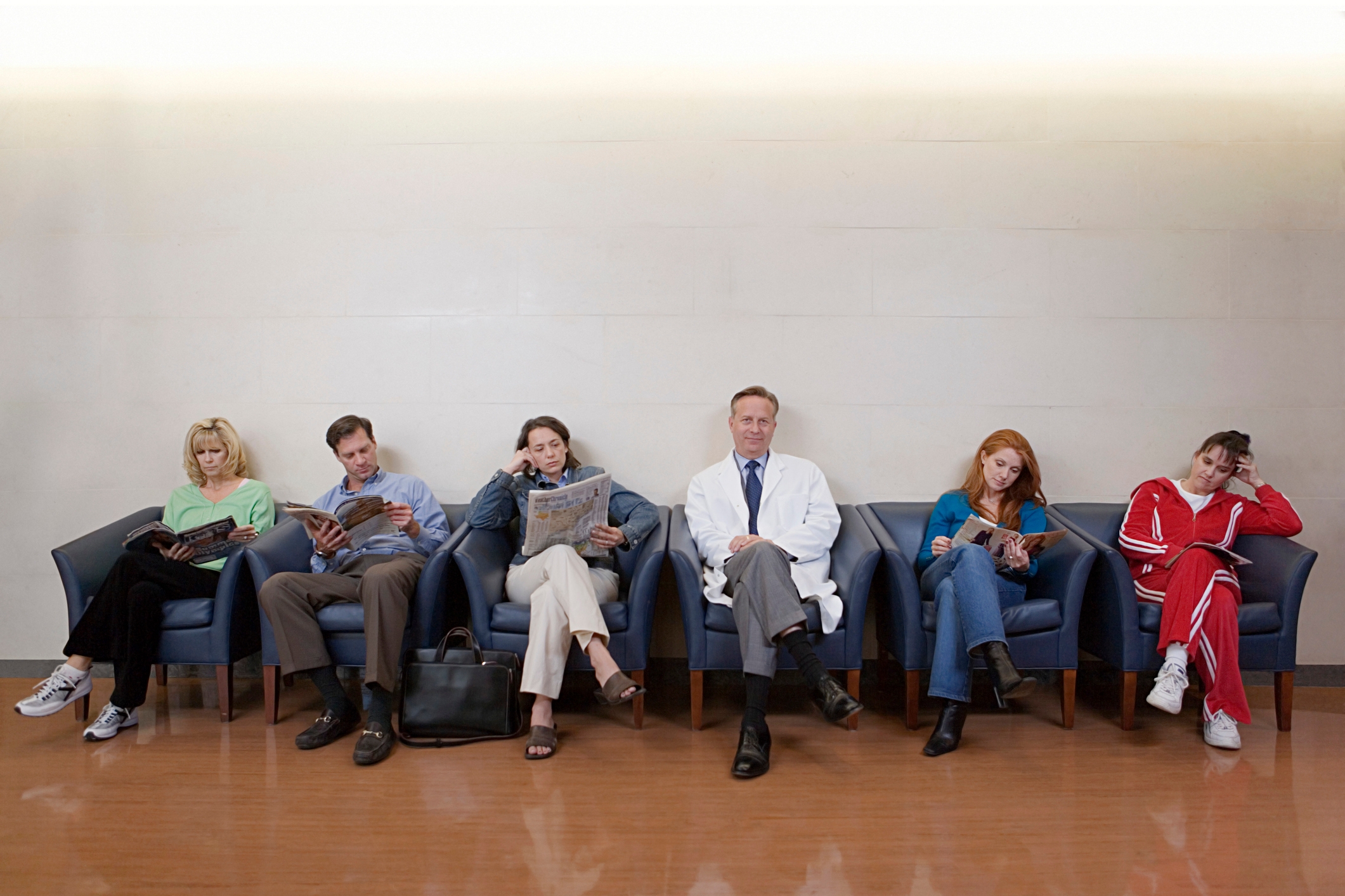 Seks personer sitter i et venteområde. En mann i en hvit frakk sitter i sentrum, med andre tilfeldig kledd, og antyder en lege