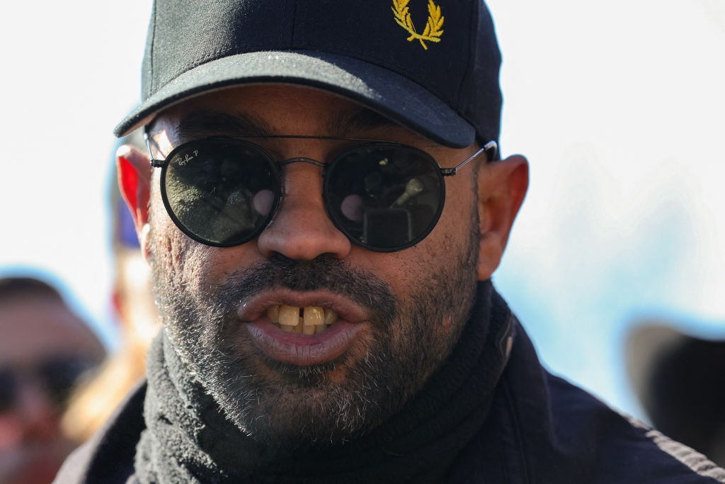 A person wearing a baseball cap, sunglasses, and a scarf, speaking outdoors