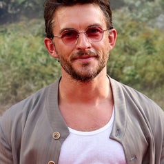 Person wearing round sunglasses, light jacket over a white tank top, outdoors with greenery in the background