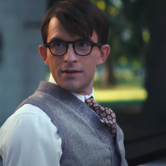 A person with glasses, wearing a vest and patterned tie, is outdoors, with a focused expression. Trees and a park setting are visible in the background