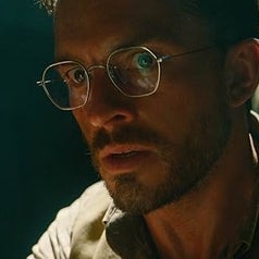 A person wearing glasses and gloves holds a syringe while looking intently forward in a dimly lit environment