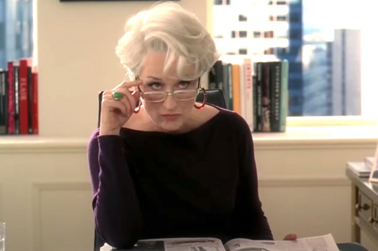Person with short, white hair, wearing glasses and a stylish top, looking over reading glasses while holding a magazine in an office setting