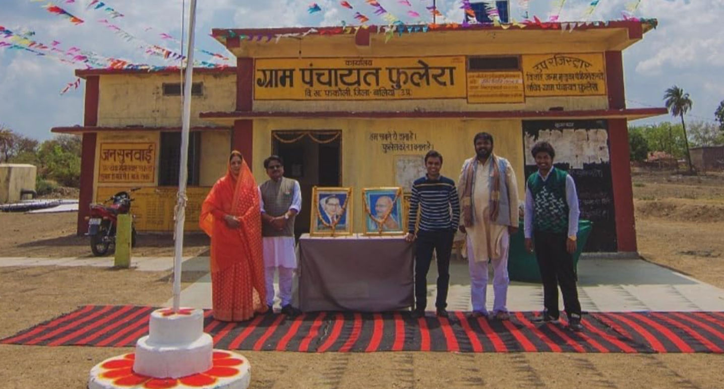 Five people stand outside a rural community building with framed photos on a table, under festive banners