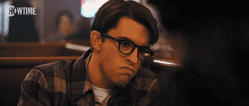 A person wearing glasses and a flannel shirt sits in a dimly lit setting, expressing a puzzled or thoughtful look