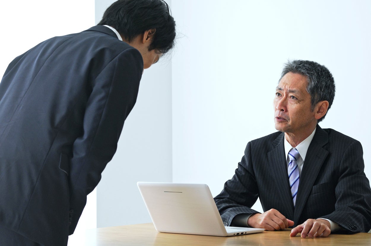 2人の男性がスーツを着てデスクに向かい合っている。1人は腰をかがめ、もう1人はノートパソコンの前で座る。ビジネスの場面。