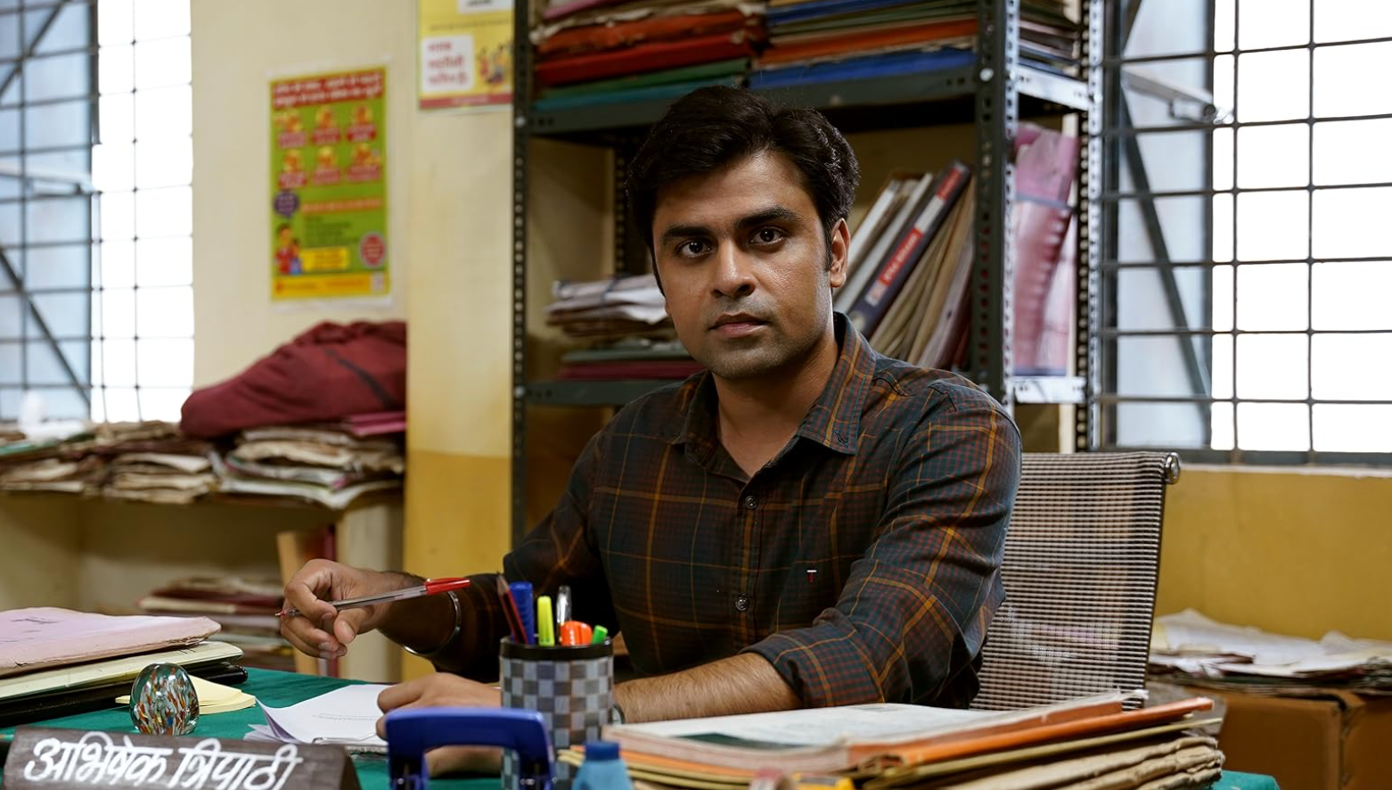 Person sitting at a cluttered office desk, holding a pen, looking directly at the camera. Shelves filled with files and paperwork in the background