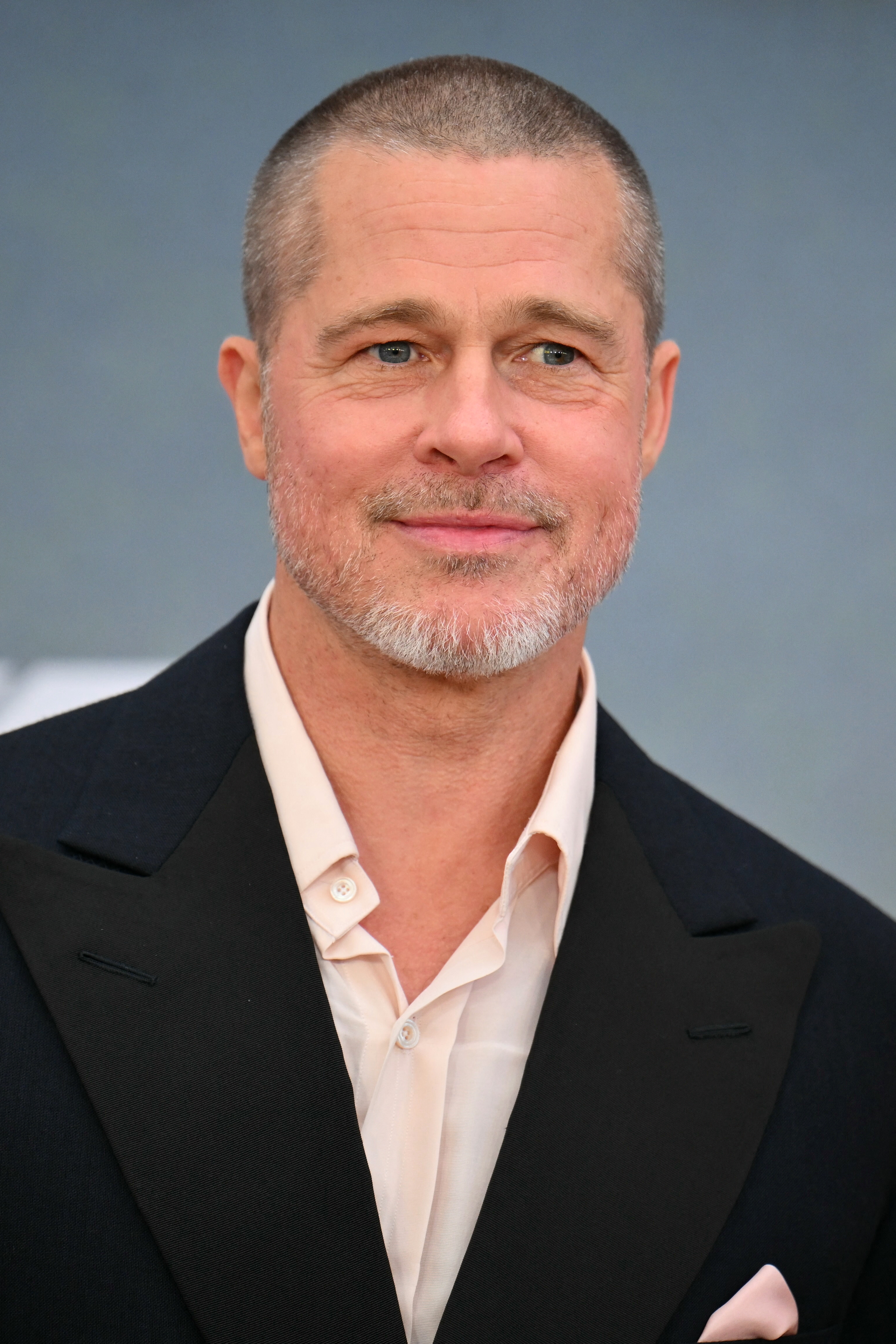 Smiling man in formal attire with a short hairstyle and light facial hair, standing against a neutral background