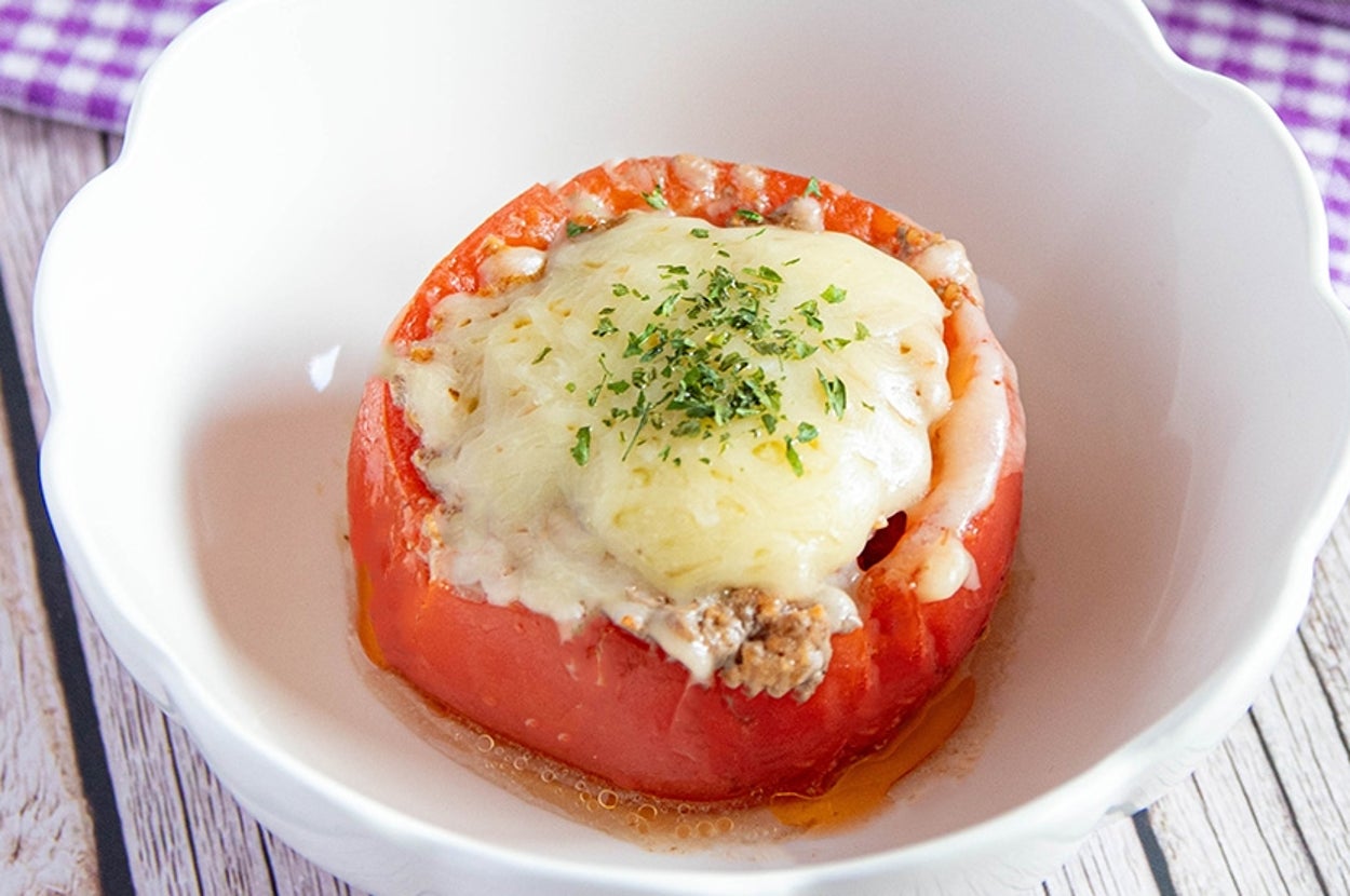 チーズとハーブがトッピングされた肉詰めトマトが白い皿に盛られています。背景に紫のテーブルクロスがあります。