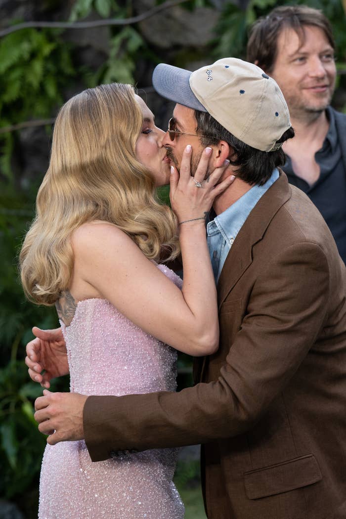 A couple kisses at an outdoor event. The woman wears a strapless sequined dress; the man is in a brown jacket and cap. Another person stands in the background