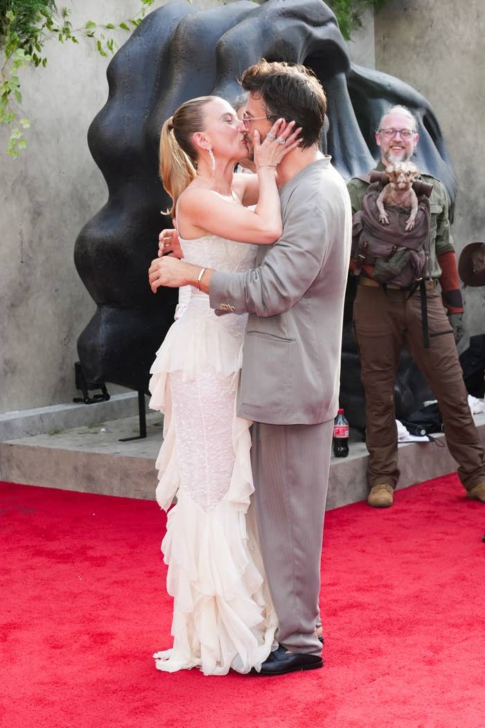 Two people embrace on a red carpet; one in a ruffled dress, the other in a suit. A bystander holds a small animal in the background