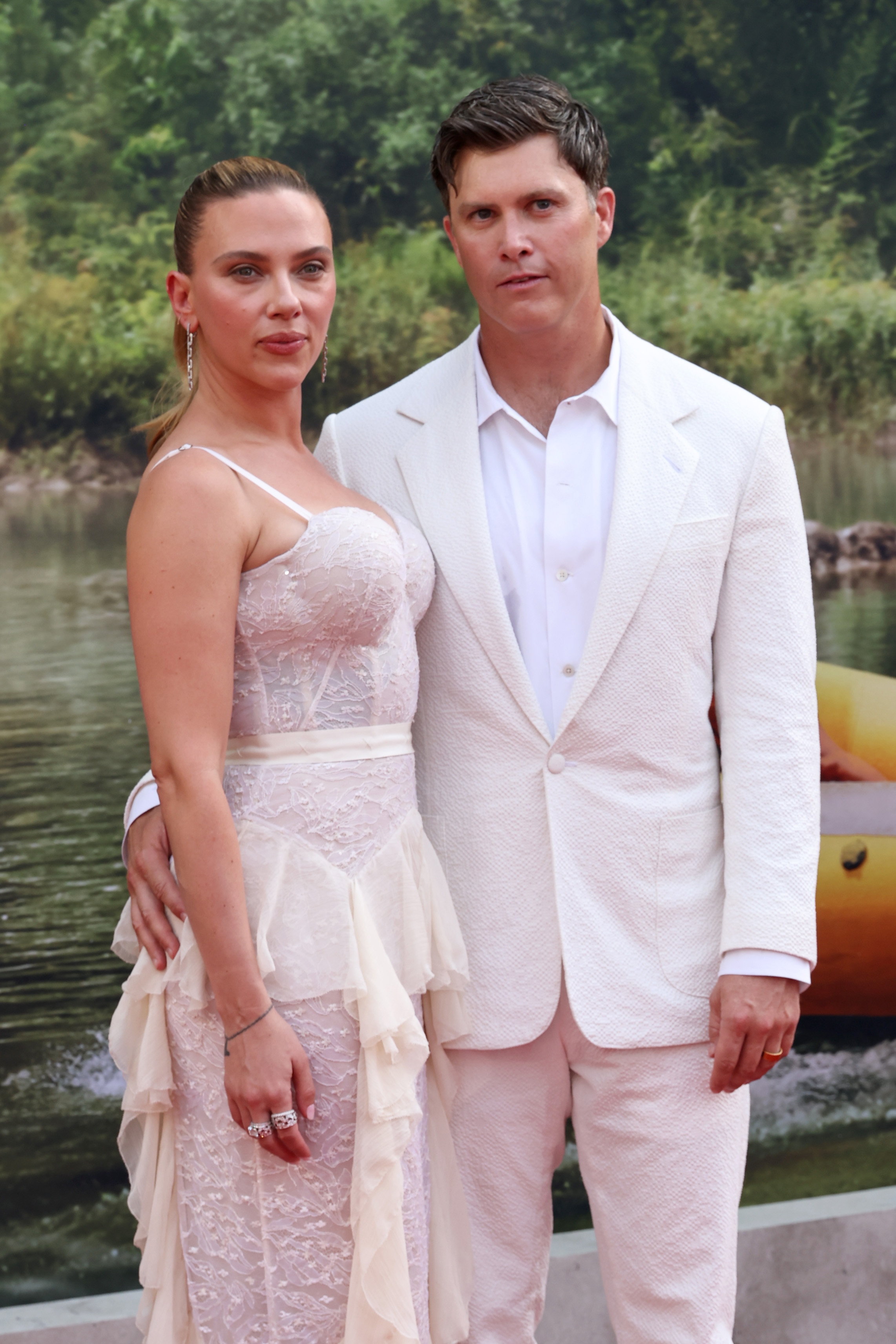 Two individuals pose outdoors. One wears a sheer lace dress; the other wears a white suit