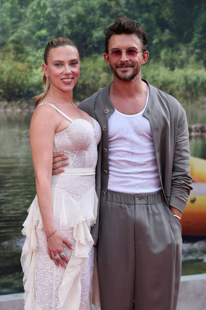 Two people pose on a red carpet. The woman wears a lacy, tiered dress. The man sports a casual suit with a white tank top and sunglasses