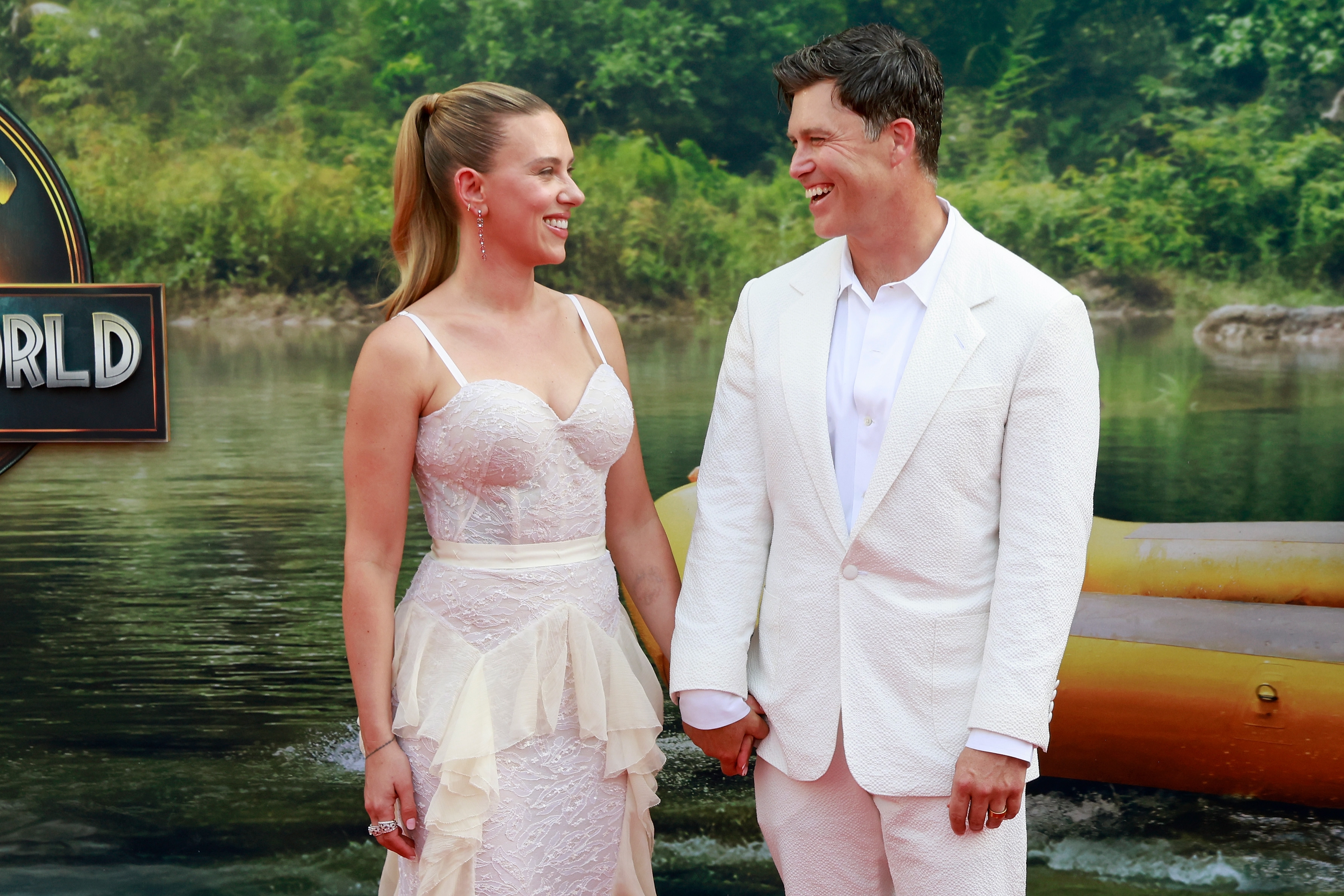 Two people smiling at each other on a red carpet. The woman wears a fitted lace dress with ruffles; the man wears a white suit. They hold hands