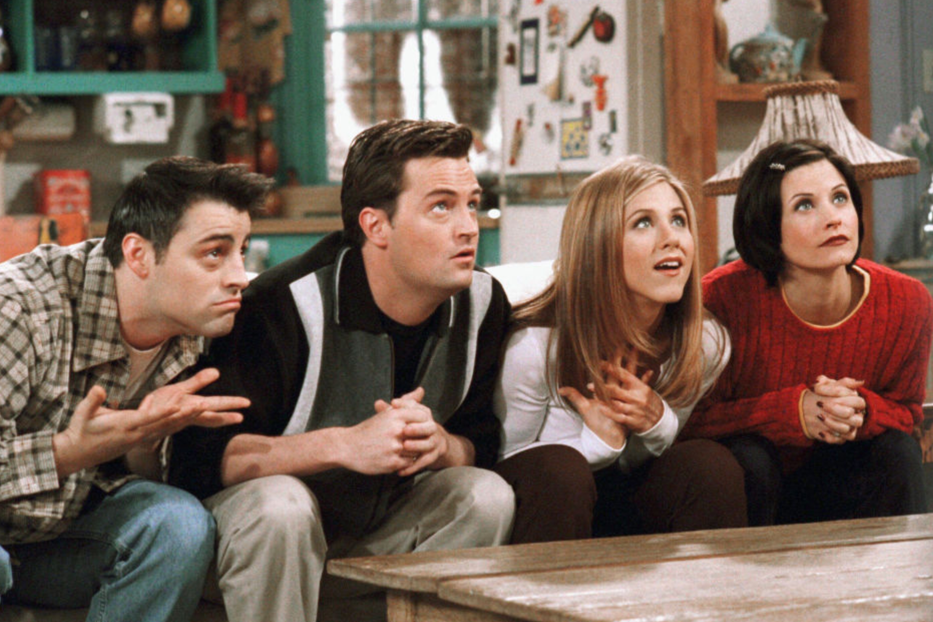 Six people sit closely on steps, casually dressed in 90s fashion