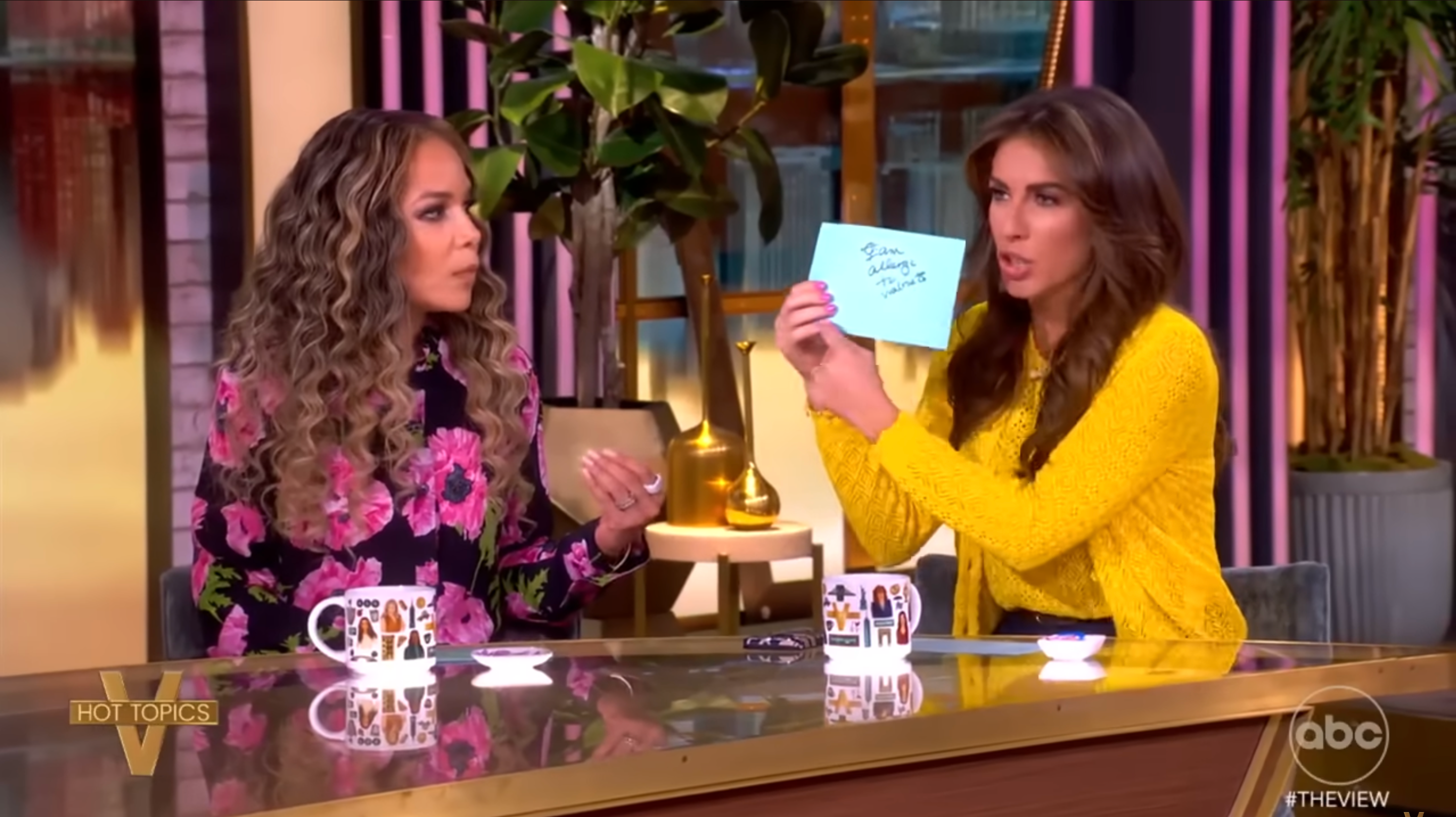 Two women sitting at a table on a talk show set, engaging in conversation. One holds a notecard with visible text