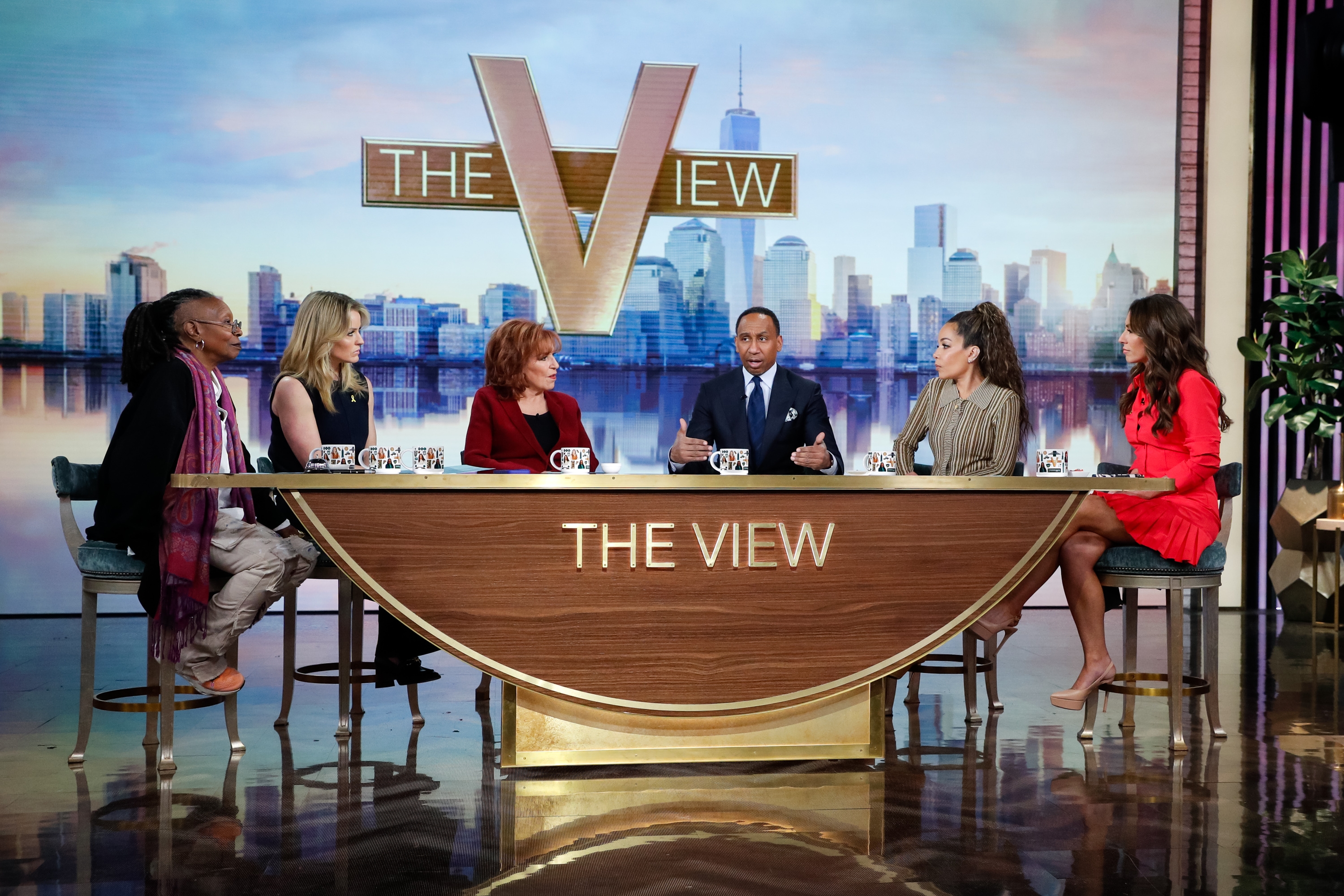 TV panel show with six hosts seated behind a desk, engaged in discussion; show's logo displayed on set
