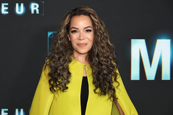 Person in elegant attire with curly hair, wearing a stylish cape-style dress, at a media event backdrop