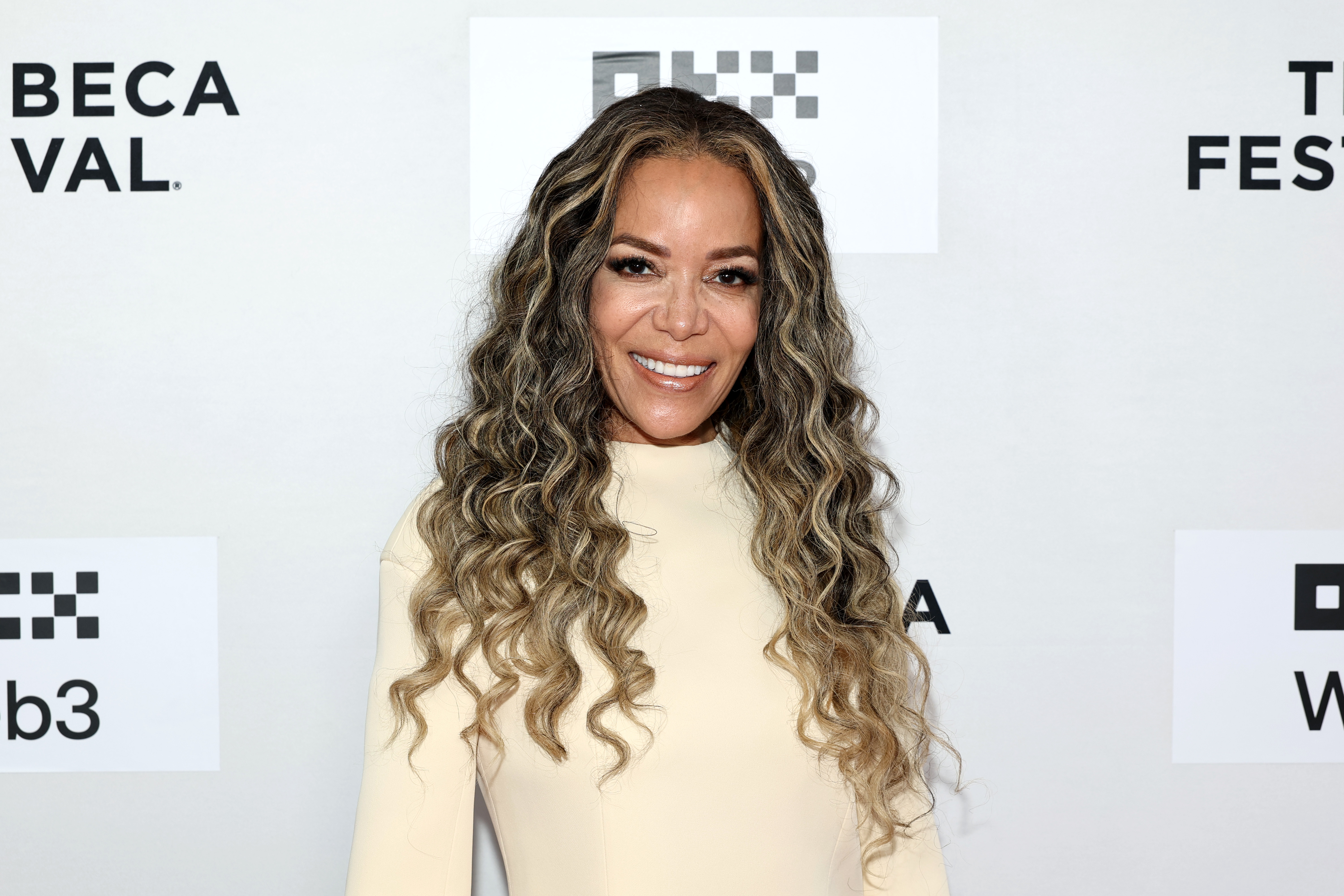 Person posing at an event, wearing a long-sleeve, knee-length dress with curly hair styled down