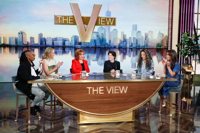 Five hosts of &quot;The View&quot; sit at a table on set, engaged in conversation. The backdrop features a city skyline