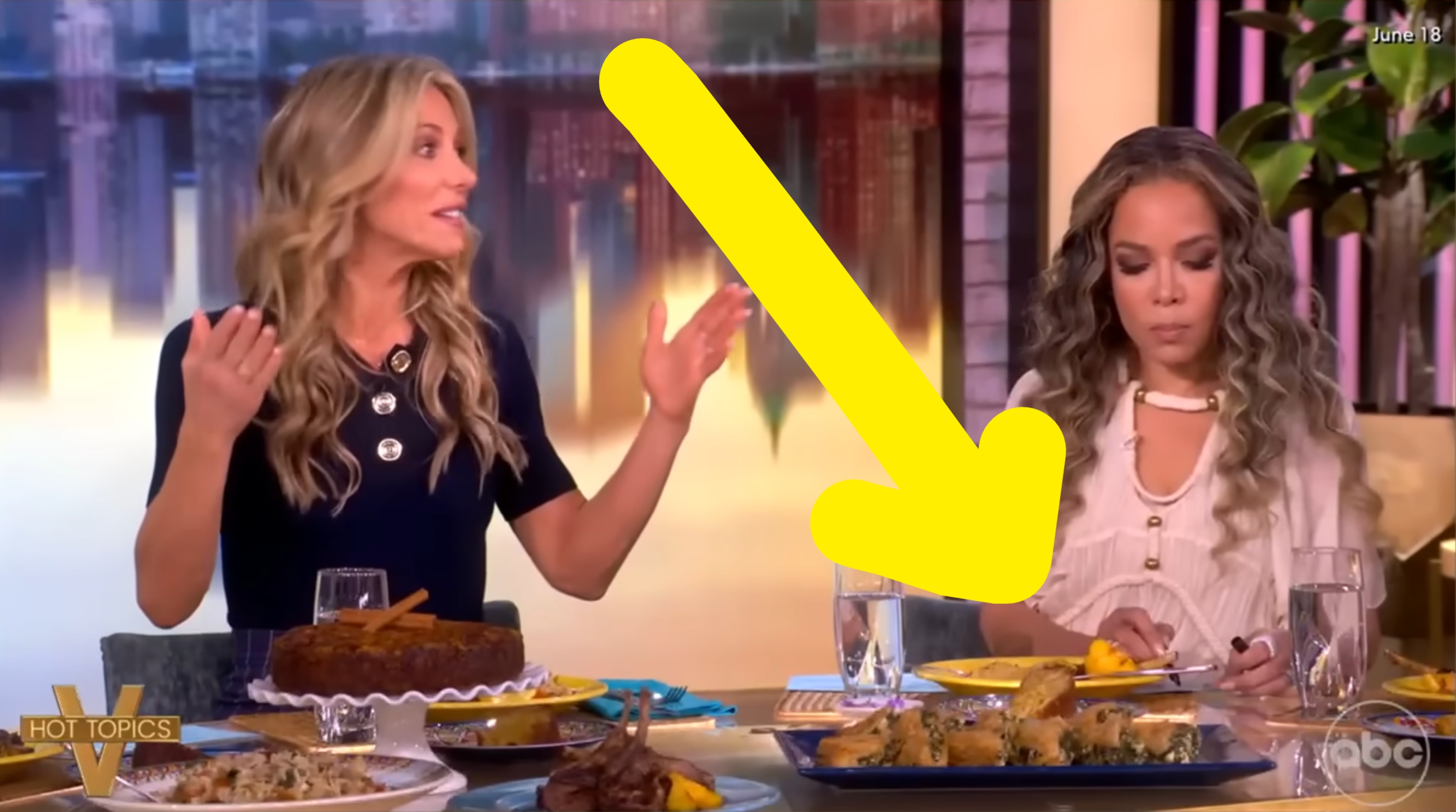 Two women on a talk show set, engaged in discussion with food on the table