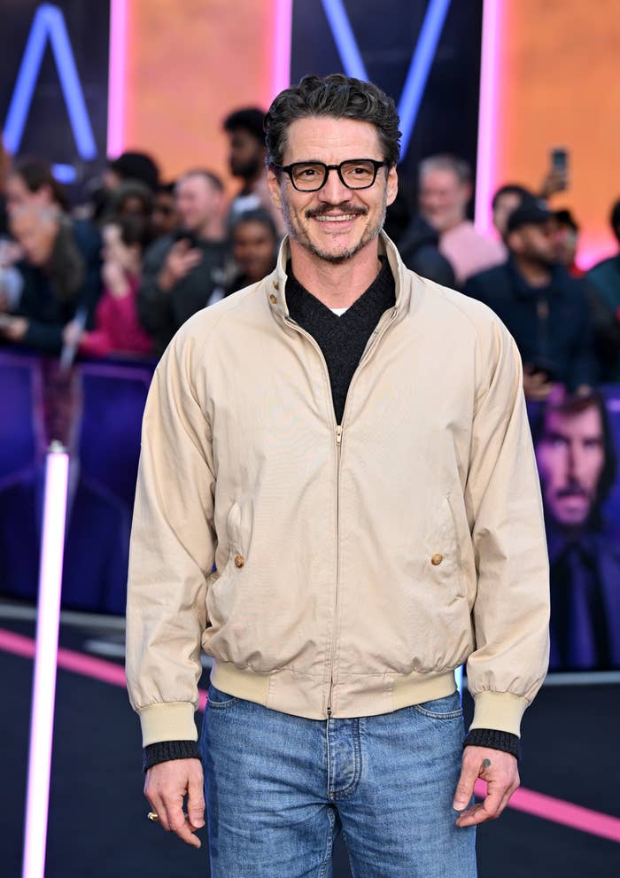 Man in casual jacket and jeans smiles on a red carpet at a celebrity event, with a backdrop of enthusiastic fans