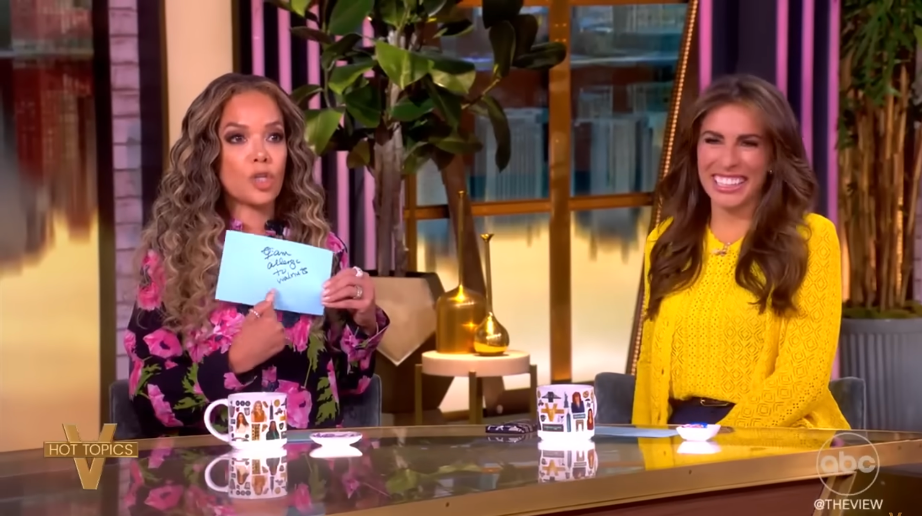 Two women sit at a talk show table. One holds a note. Both are smiling, and there's a plant in the background