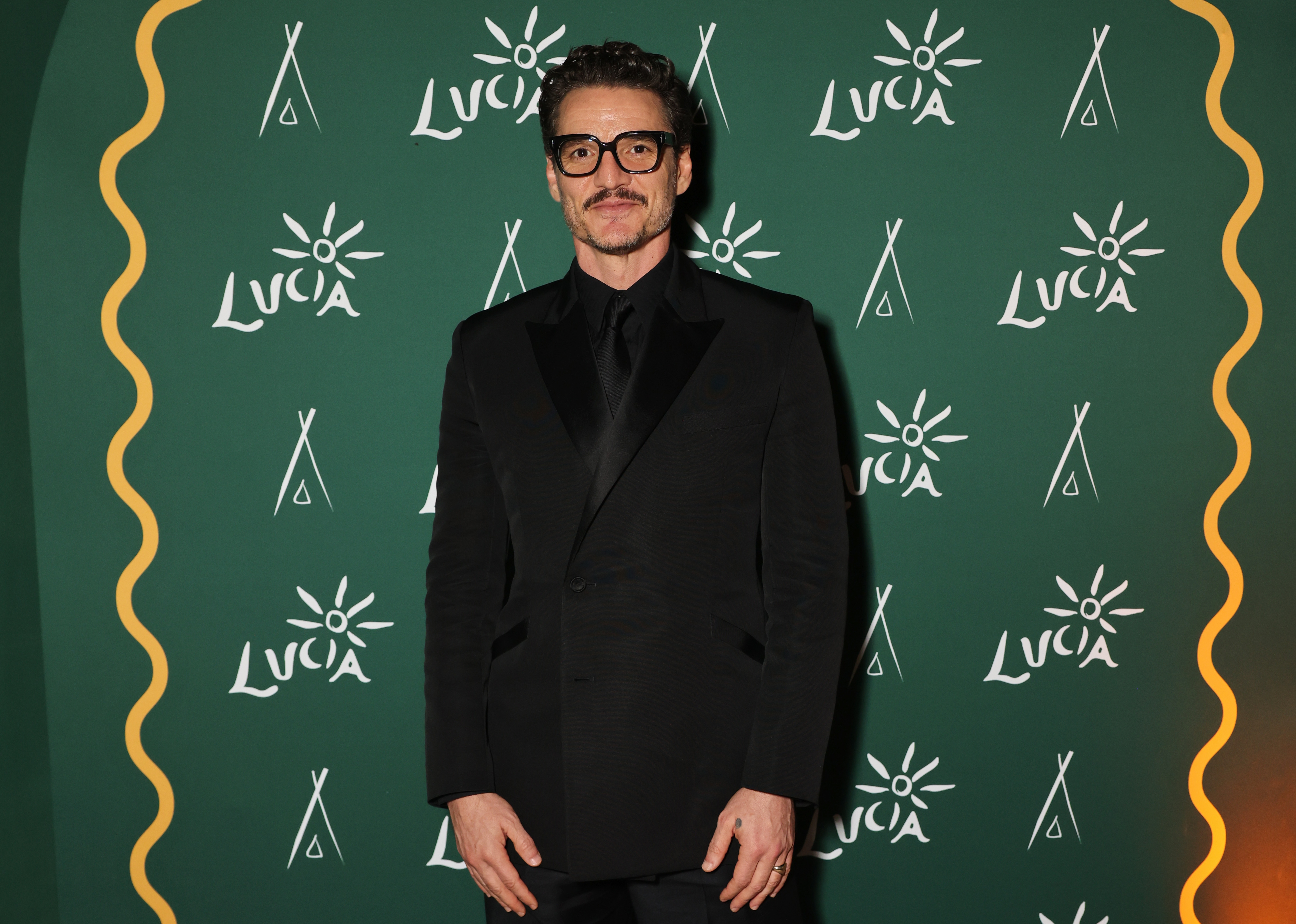 Person in elegant black suit with glasses at a red carpet event, posing in front of a green backdrop with "Luca" logos