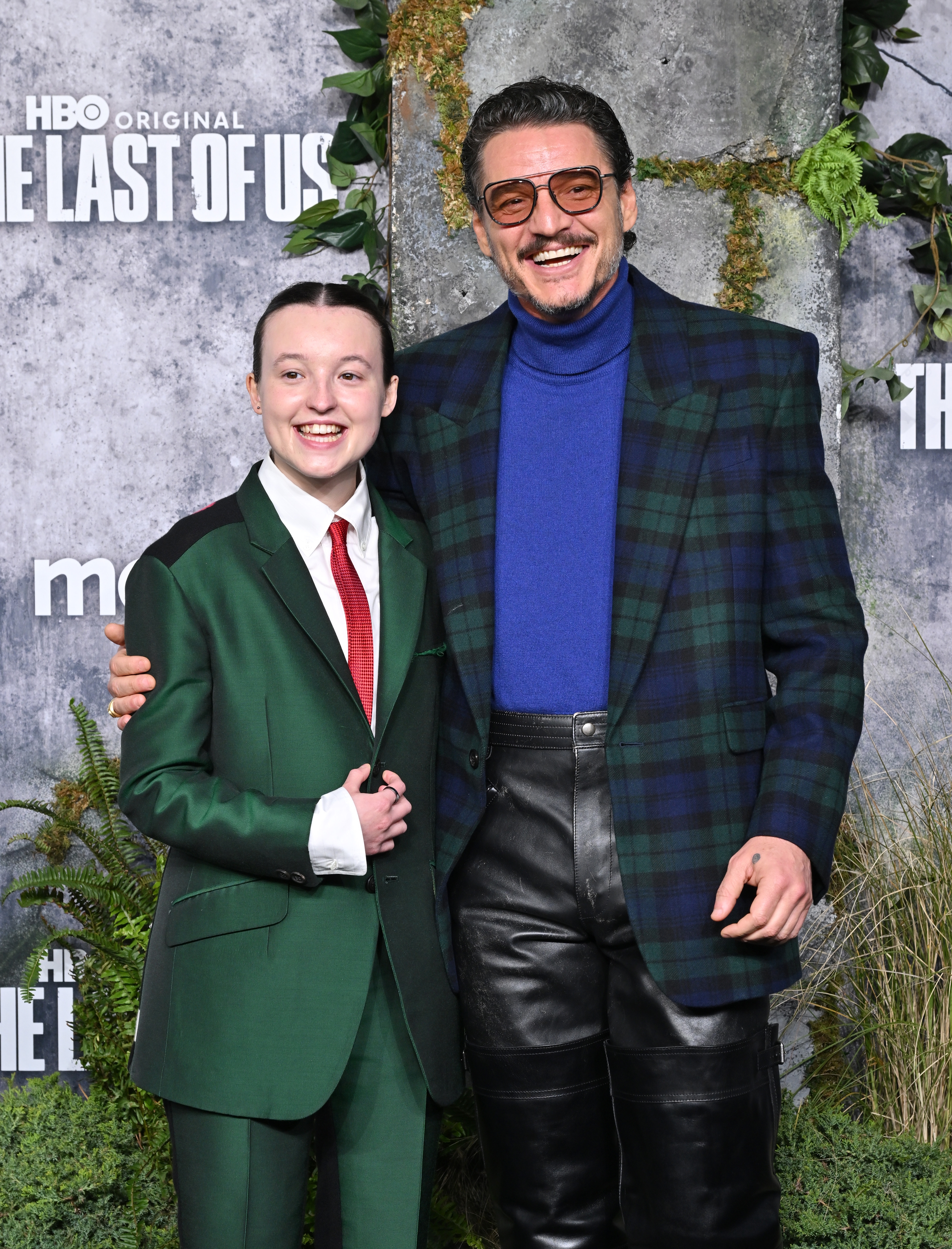 Two individuals on a red carpet; one in a green suit with a tie, another in a plaid blazer and turtleneck, both smiling warmly at the event