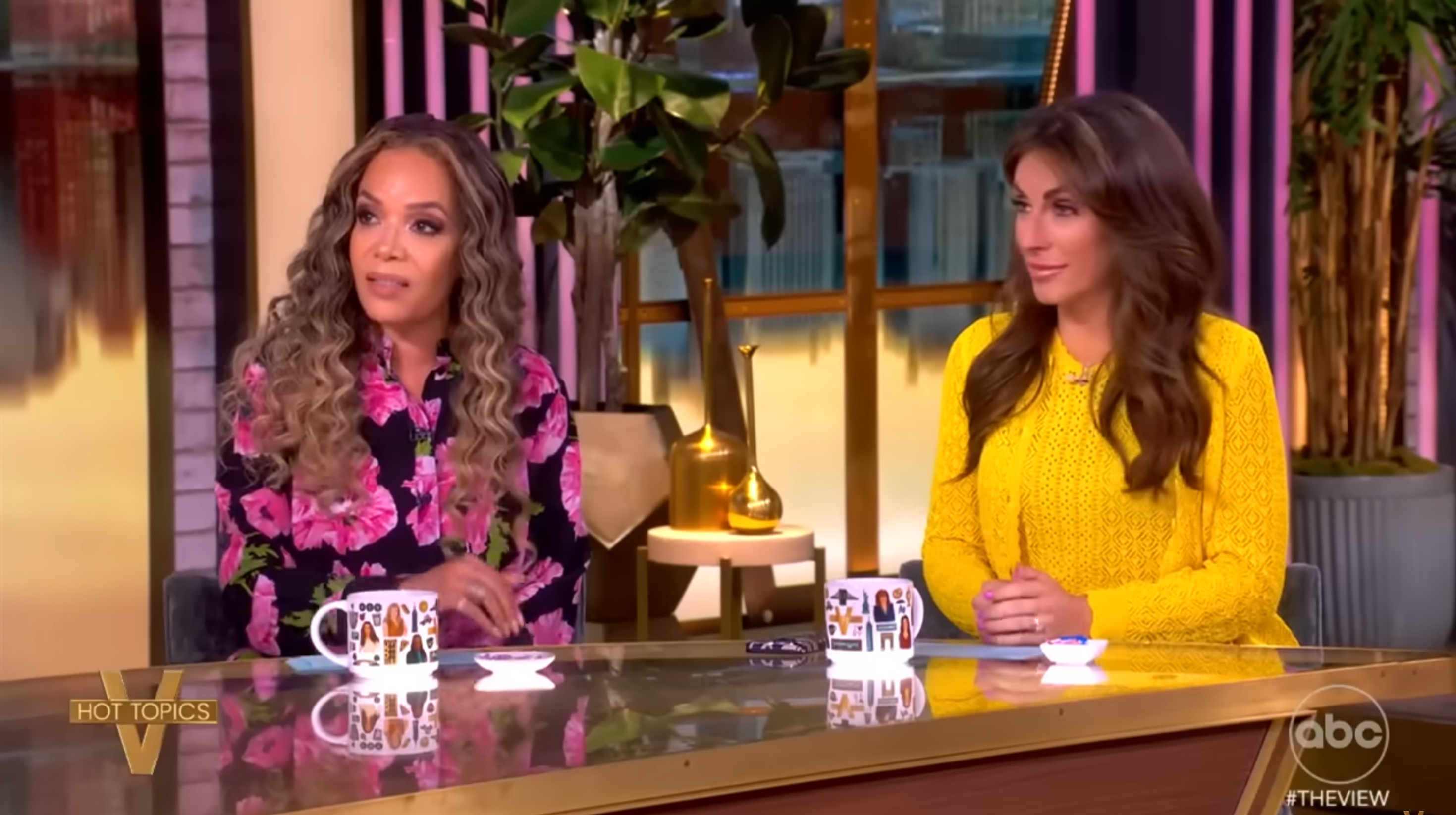 Two individuals sit at a talk show desk. One wears a floral print outfit, while the other is in a textured outfit. They appear engaged in discussion