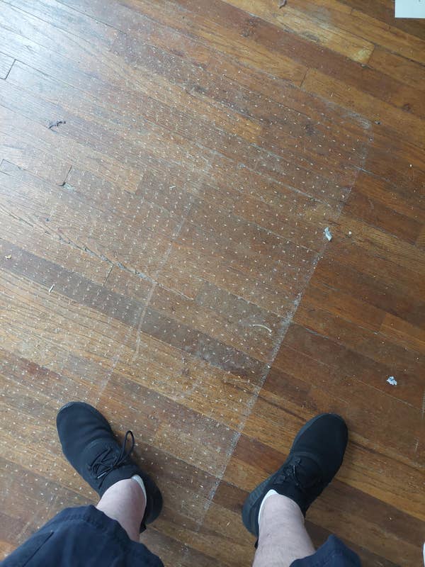 A person wearing black sneakers stands on a wood floor with a patch of worn-out or missing finish