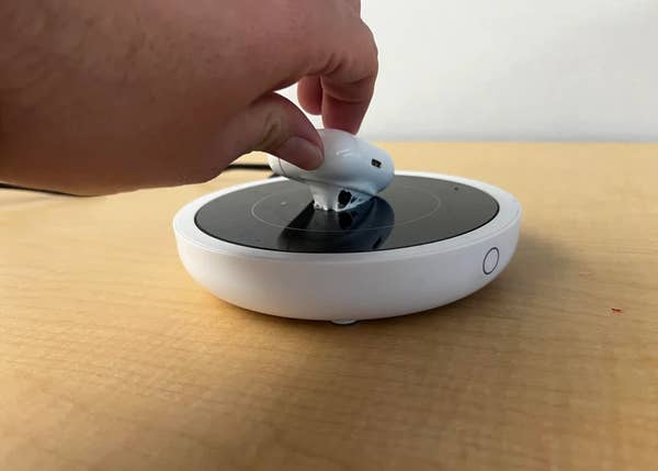 A hand places a white earbud onto a circular wireless charger resting on a wooden surface