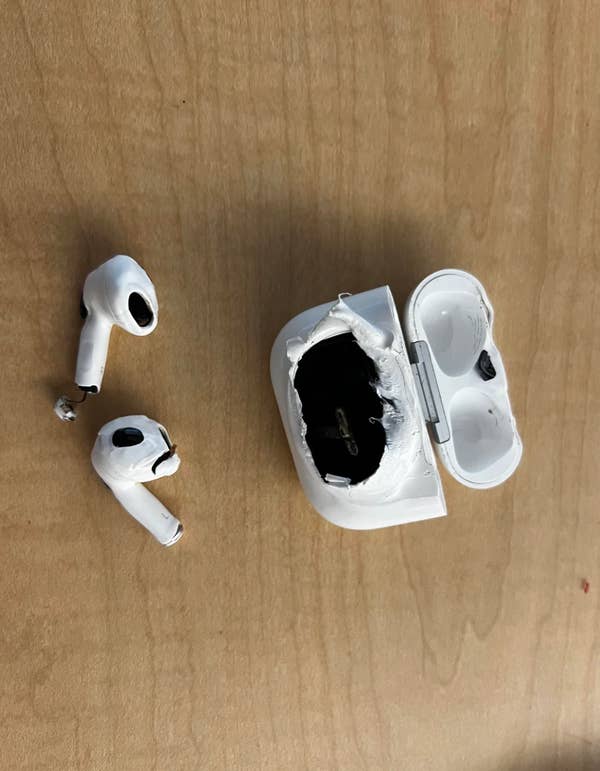 Damaged wireless earbuds with their charging case. The earbuds and case appear melted or burned, showing significant wear and damage