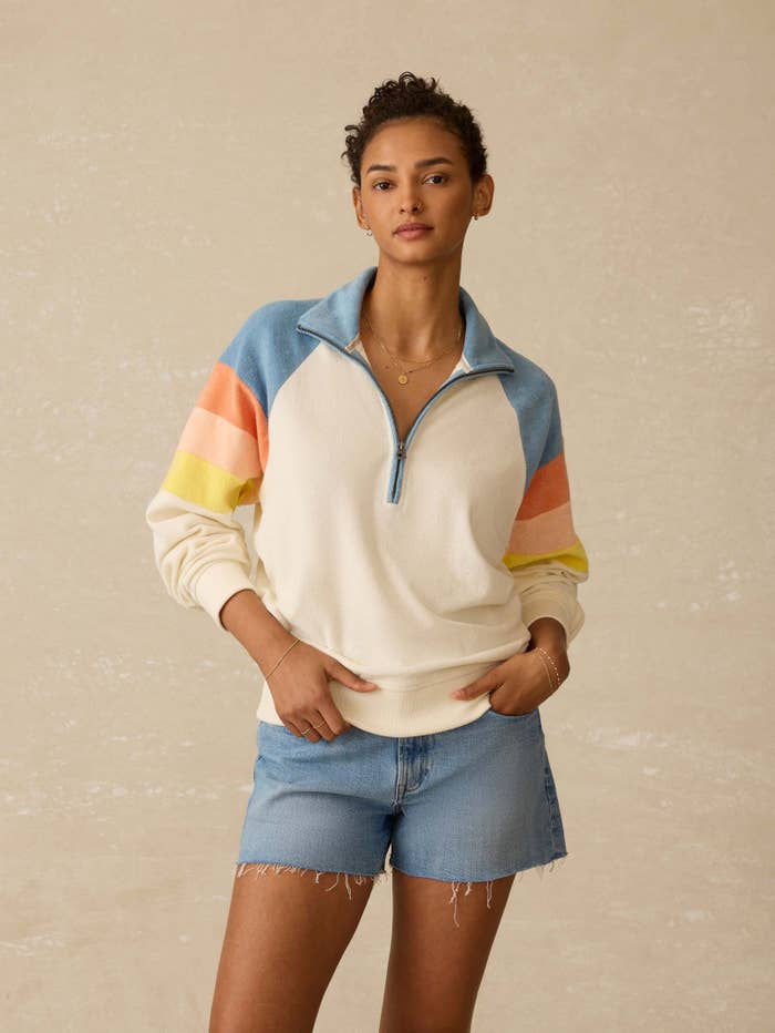 model wearing a casual, zip-up sweater with colorful stripes connected  the sleeves and denim shorts