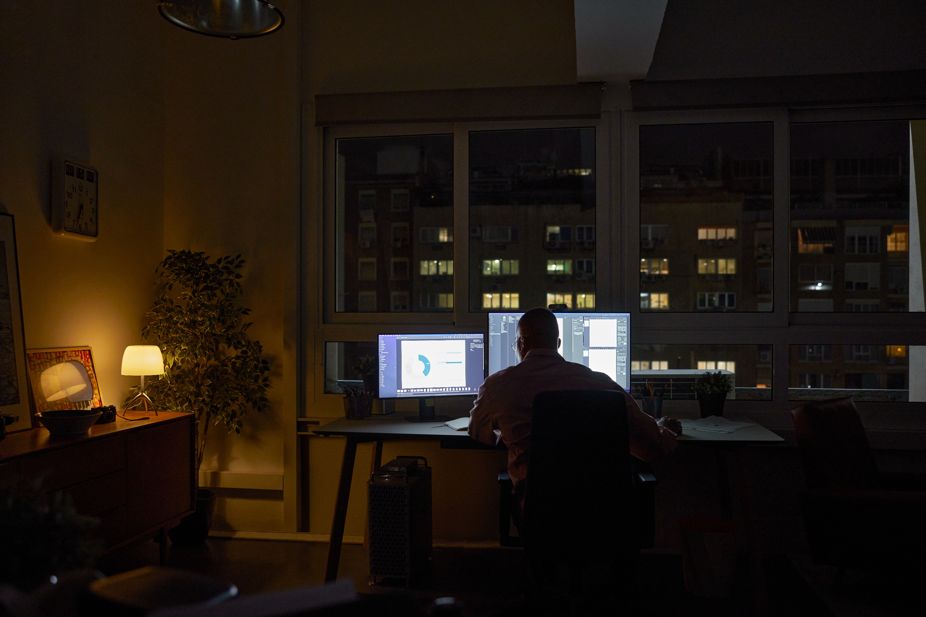 Person som jobber med doble skjermer i et svakt opplyst rom med utsikt over bybygg om natten, og skaper et fokusert og produktivt miljø