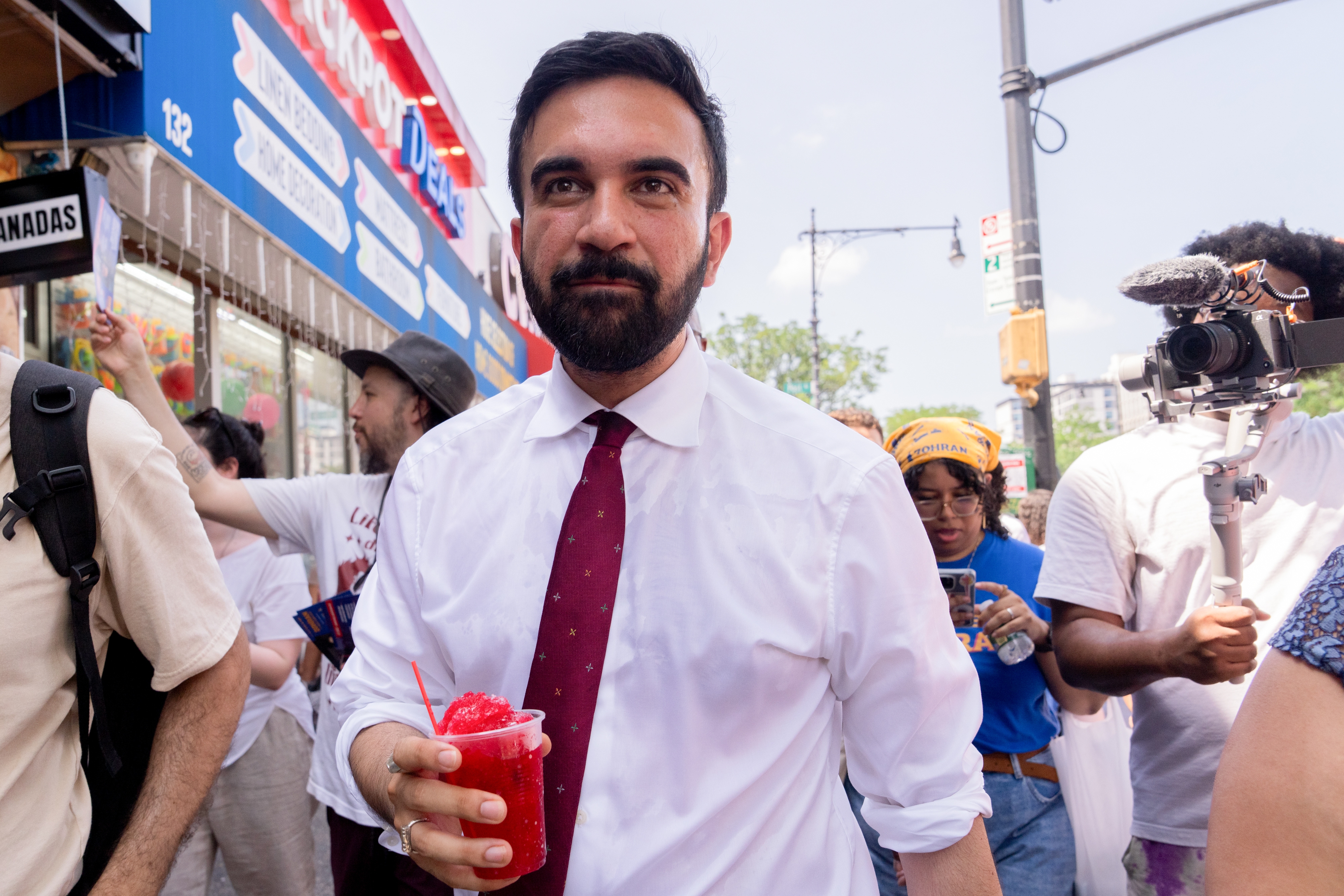 Person in white shirt and red tie holds a red drink while walking outdoors, surrounded by people, some filming the scene